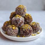 A stack of rich, dark Dubai chocolate balls rolled in shredded coconut and topped with crushed pistachios.