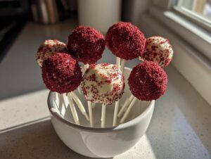 A cluster of homemade red velvet cake pops, some fully coated in red crumbs and others dipped in white chocolate, standing in a small white bowl.