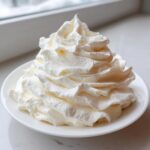 A tall swirl of rich, fluffy vanilla buttercream frosting piled high on a small white plate.