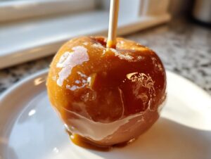 A close-up of a glossy, homemade caramel apple on a stick, resting on a white plate.