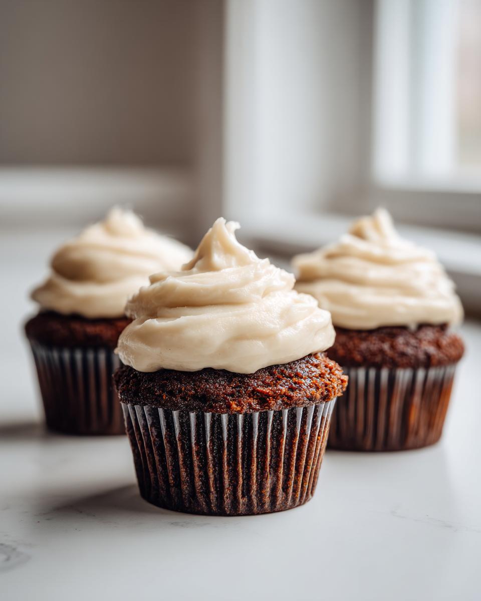 Three dark, rich gingerbread cupcakes topped with creamy, light-colored frosting sitting on a white marble surface.