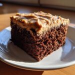 A moist, dark slice of gingerbread cake topped with thick, creamy brown frosting, sitting on a white plate.