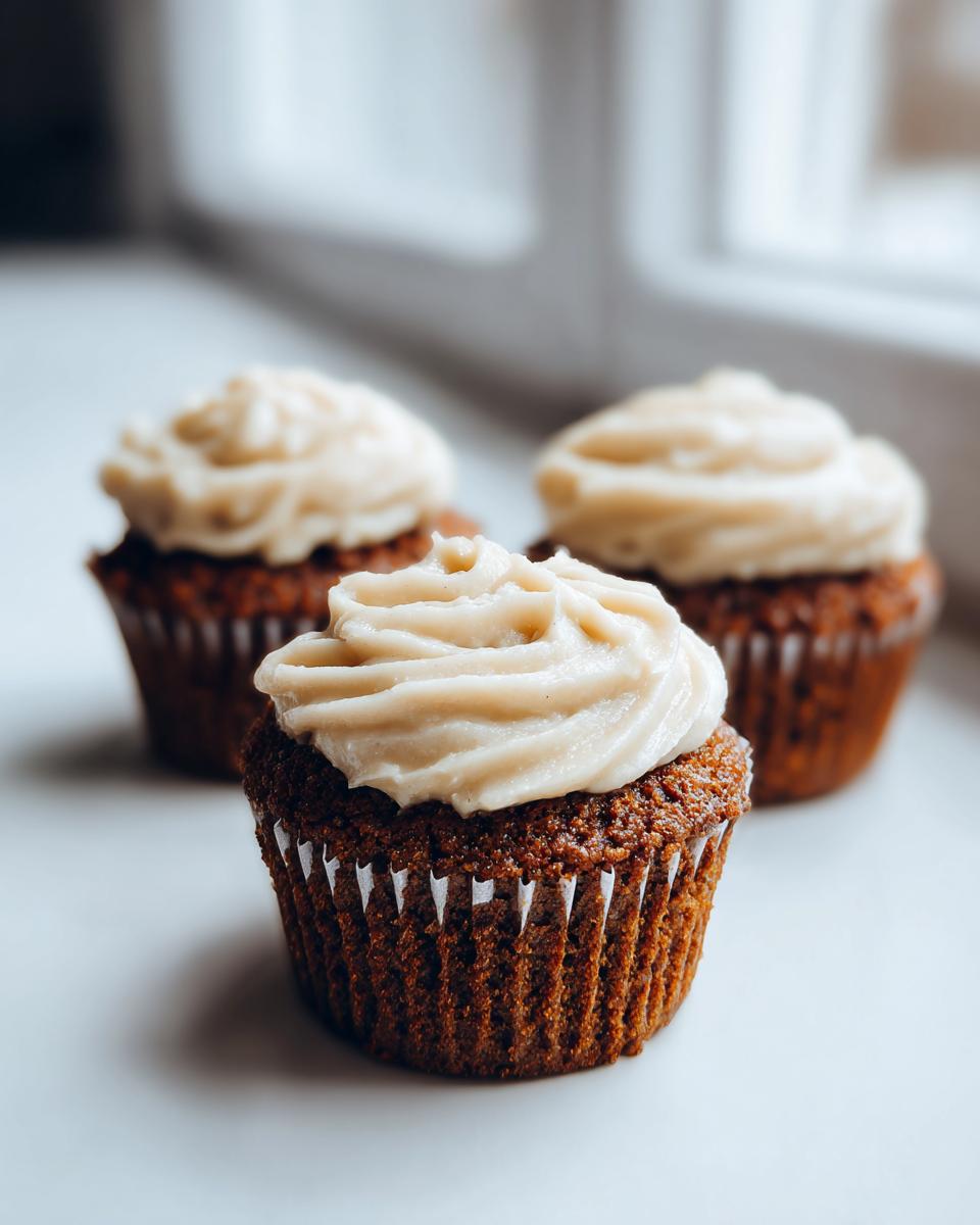Three dark brown gingerbread cupcakes topped with creamy, pale frosting, sitting on a white surface near a window.