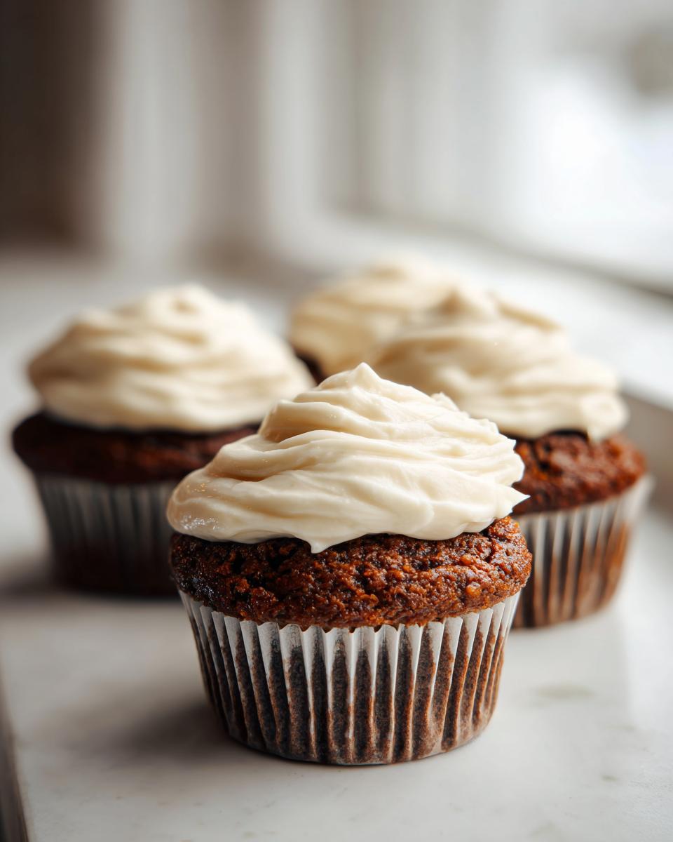 Three rich, dark gingerbread cupcakes topped with thick swirls of cream cheese frosting, sitting near a window.