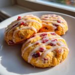 Three golden cranberry orange cookies drizzled with white glaze, sitting on a white plate near a window.