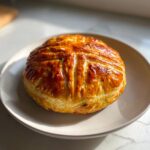 A perfectly golden brown baked brie in puff pastry, scored on top, resting on a light-colored plate.