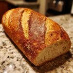 A freshly baked loaf of french bread with a deeply golden, scored crust resting on a granite countertop.