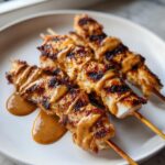 Four skewers of perfectly grilled chicken satay drizzled generously with creamy peanut sauce on a white plate.