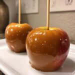 Close-up of two shiny, homemade caramel apples on sticks resting on a white plate.