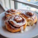 Three gooey, freshly baked christmas tree cinnamon rolls covered in thick white icing on a white plate.