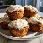 A stack of moist carrot cake muffins generously topped with thick cream cheese frosting.
