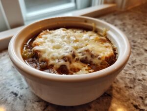 A close-up of a bowl of rich, dark philly cheese steak soup topped with a thick layer of melted, bubbly white cheese.