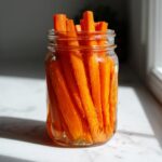 Bright orange carrot sticks packed tightly in a clear glass jar, ready to be enjoyed as pickled carrots.