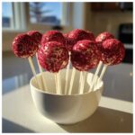 Close-up of several white-dipped red velvet cake pops covered in red sprinkles, displayed in a white bowl.