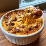 A spoonful of hot, cheesy sausage cream cheese dip being lifted from a white ramekin, showing a cheese pull.
