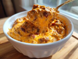 A spoonful of hot, cheesy sausage cream cheese dip being lifted from a white bowl, showing a dramatic cheese pull.