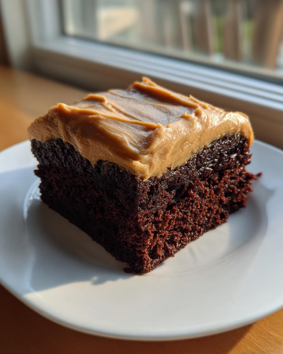 A close-up of a moist, dark slice of gingerbread cake topped with thick, light brown caramel frosting, sitting on a white plate.