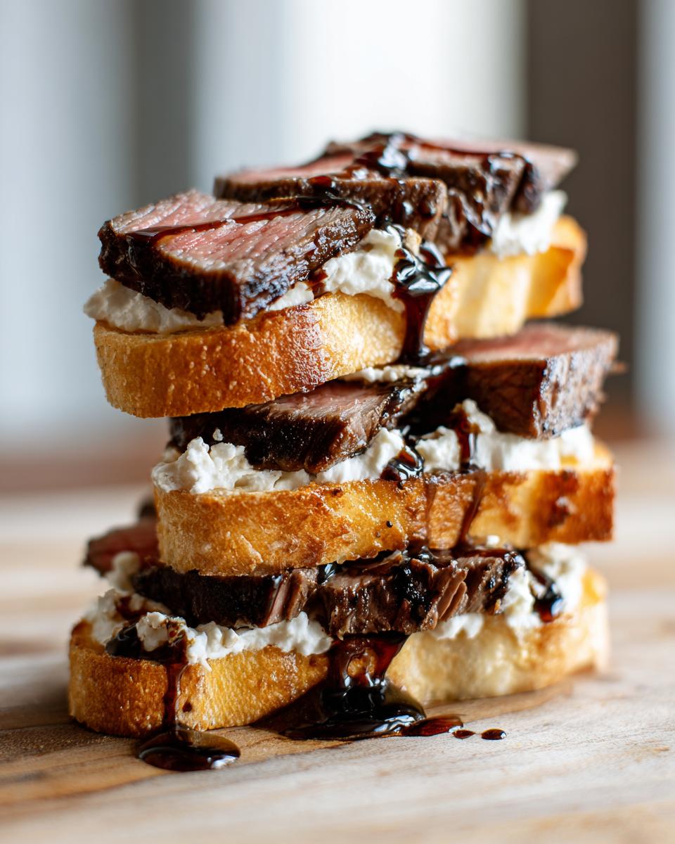 A tall stack of steak crostini featuring seared steak, creamy cheese, and a dark balsamic glaze drizzle.