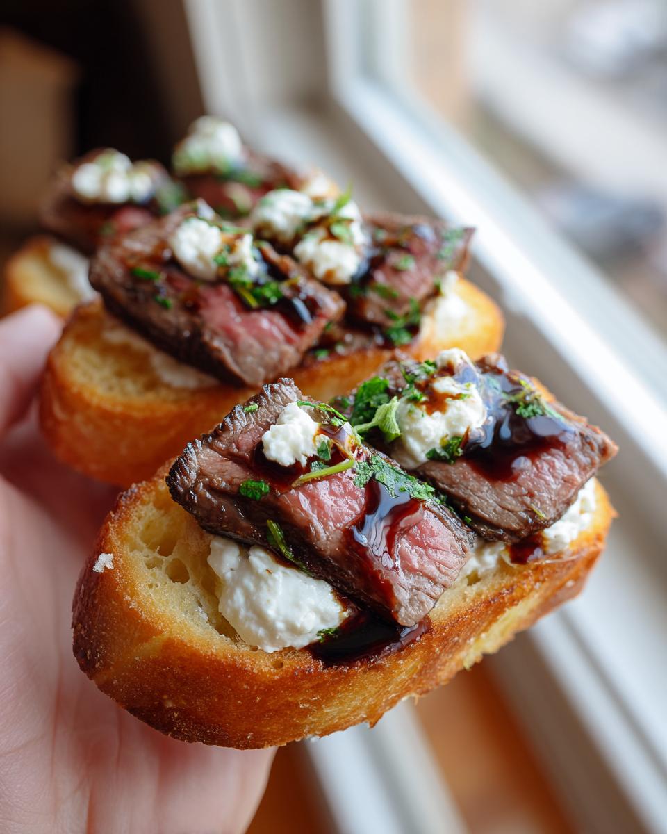 Close-up of a hand holding a piece of steak crostini topped with medium-rare steak, creamy cheese, and balsamic glaze.