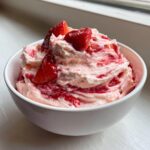 Close-up of a white bowl filled with swirled pink and white strawberry cheesecake dip, topped with fresh strawberry slices.