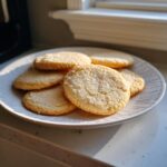 A plate piled high with freshly baked, slightly crackled vegan sugar cookies dusted with sugar.