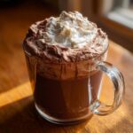A clear glass mug filled with rich, dark whipped hot chocolate topped generously with whipped cream and a dusting of cocoa.