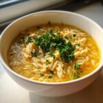 Close-up of a white bowl filled with hot chicken orzo soup, garnished generously with fresh parsley.