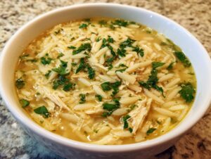 A close-up view of a bowl of amazing chicken orzo soup, featuring shredded chicken, orzo pasta, and fresh parsley garnish.