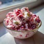 A close-up of a fluffy, pink cranberry salad mixed with whipped cream and chunks of fruit in a clear glass bowl.
