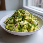 A white bowl filled with vibrant green avocado egg salad, featuring chunks of hard-boiled egg and red onion, topped with fresh dill.