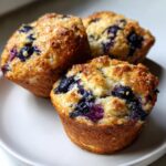 Three golden-brown blueberry cottage cheese muffins with a sparkling sugar crust served on a white plate.