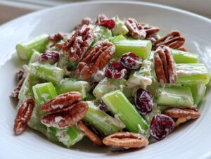 Close-up of a creamy celery salad featuring bright green celery sticks, whole pecans, and dried cranberries in a white bowl.