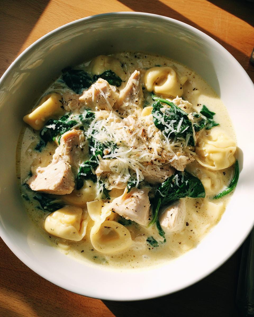 A white bowl filled with creamy chicken tortellini soup, featuring chunks of chicken, wilted spinach, and grated Parmesan cheese.