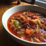 Close-up of a bowl of rich, red cabbage soup filled with ground beef, cabbage chunks, and diced carrots.