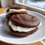 Two rich chocolate whoopie pies filled with thick white cream frosting sitting on a white plate.