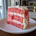 A tall, moist slice of pink strawberry cake layered with white cream cheese frosting on a white plate.