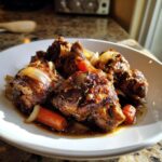 Close-up of rich, dark brown stew chicken pieces served with caramelized onions and carrots.