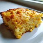 A close-up of a golden slice of moist jiffy corn casserole studded with whole corn kernels on a white plate.