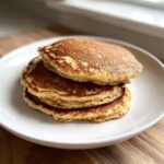 A close-up of a stack of three golden brown oatmeal pancakes served on a white plate.