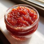 A close-up, sunlit view of rich, textured strawberry freezer jam filling a small glass jar.