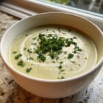 A close-up of a bowl of creamy celery soup, swirled with cream and topped with fresh chopped parsley.