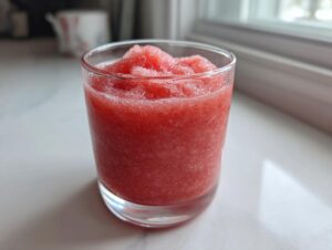 Close-up of a vibrant, icy watermelon smoothie served in a clear glass on a white surface.