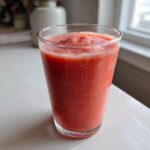 A tall glass filled with a vibrant, icy watermelon smoothie sitting on a white countertop.