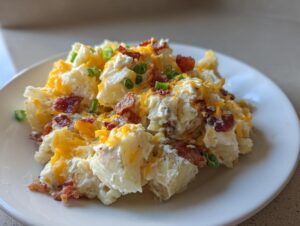 A close-up of a serving of amazing baked potato salad topped with shredded cheddar, bacon bits, and green onions on a white plate.