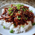 Close-up of shredded crockpot teriyaki chicken coated in glossy sauce, served over white rice and topped with green onions and sesame seeds.