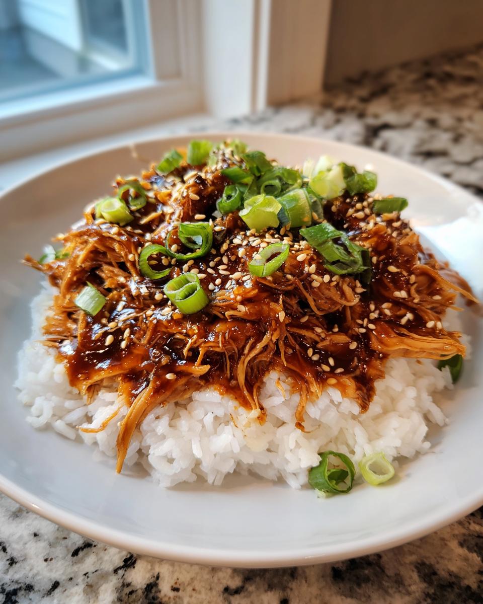 A bowl of shredded crockpot teriyaki chicken glazed with sauce, served over white rice and topped with sesame seeds and green onions.