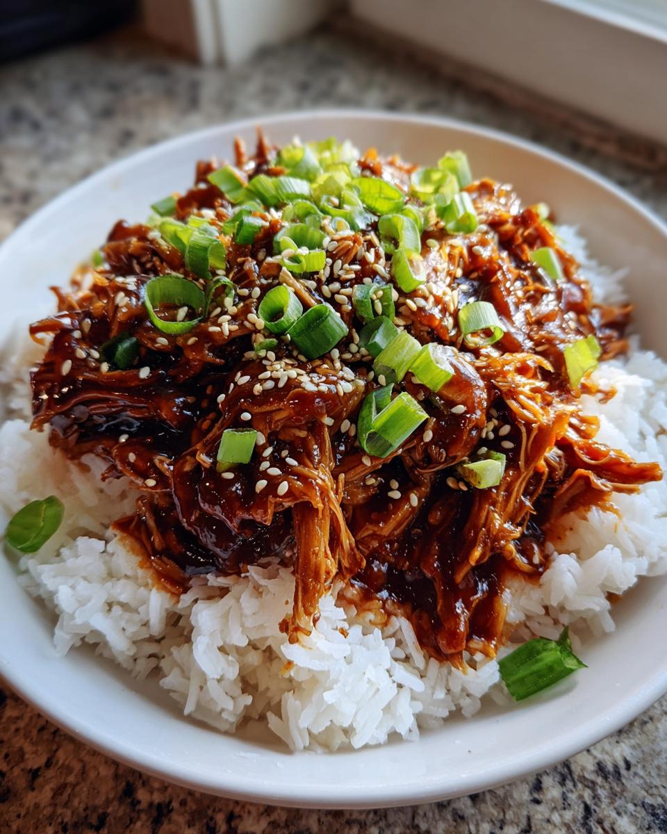 A bowl of shredded crockpot teriyaki chicken glazed with sauce, served over white rice and topped with green onions and sesame seeds.