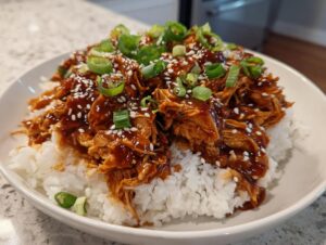 A close-up of shredded crockpot teriyaki chicken coated in a rich sauce, served over white rice and topped with sesame seeds and green onions.