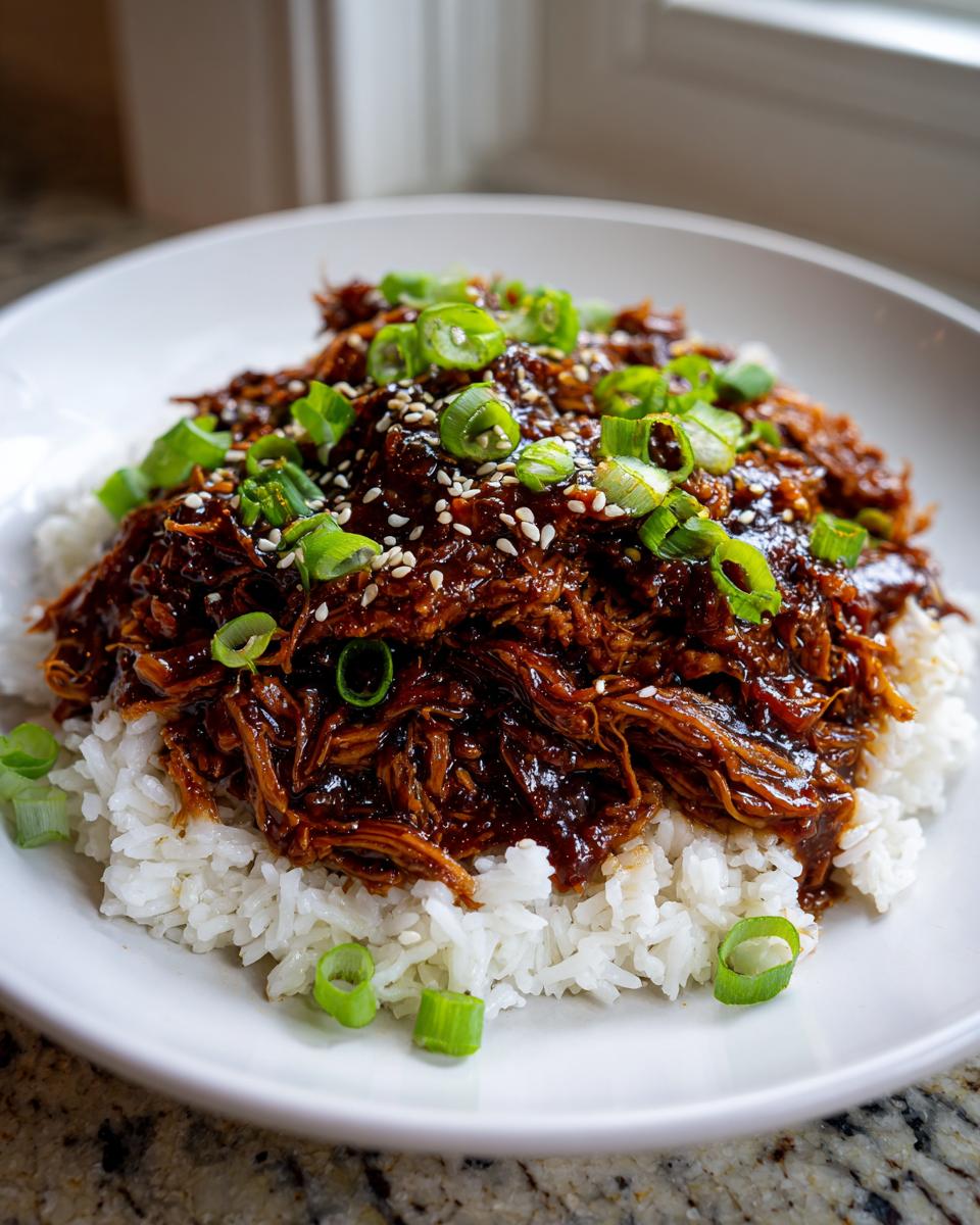 Shredded crockpot teriyaki chicken coated in dark sauce, served over white rice and topped with green onions and sesame seeds.