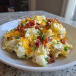 A bowl of loaded baked potato salad topped with melted cheddar cheese, crispy bacon bits, and green onions.